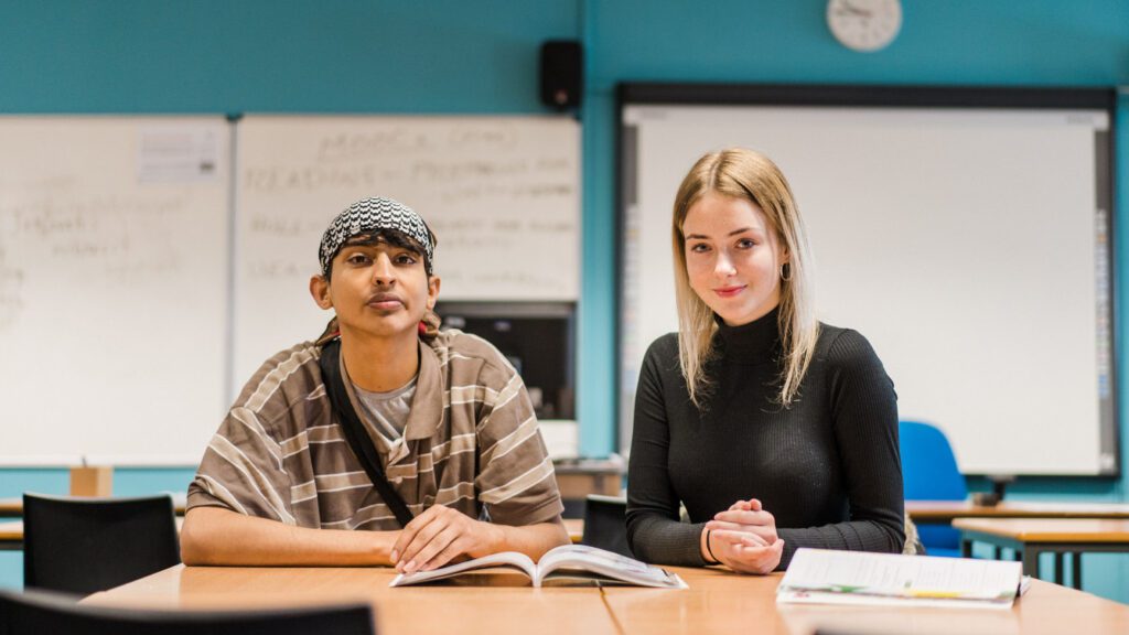 Male and female student sat at a table looking at the camera